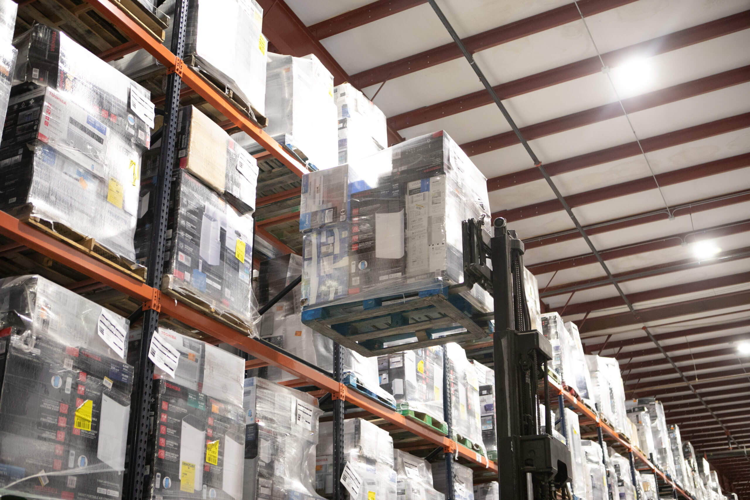 A third party logistics provider is using a forklift to raise a pallet of goods onto an upper shelf in a warehouse